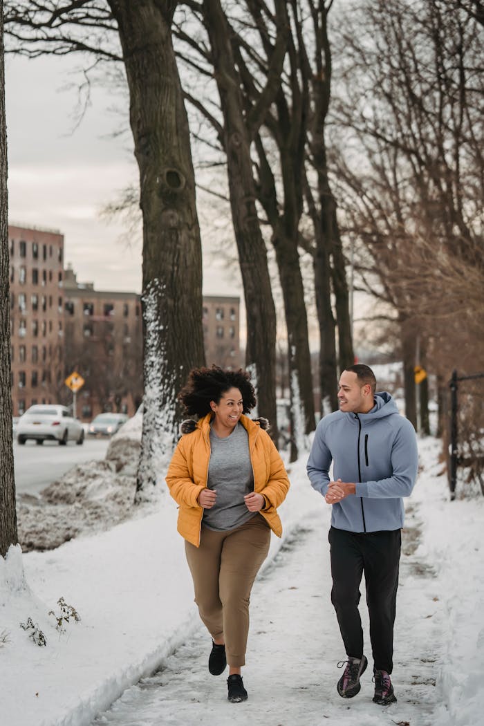 Two people jogging in winter attire on a snowy sidewalk lined with trees in an urban area.